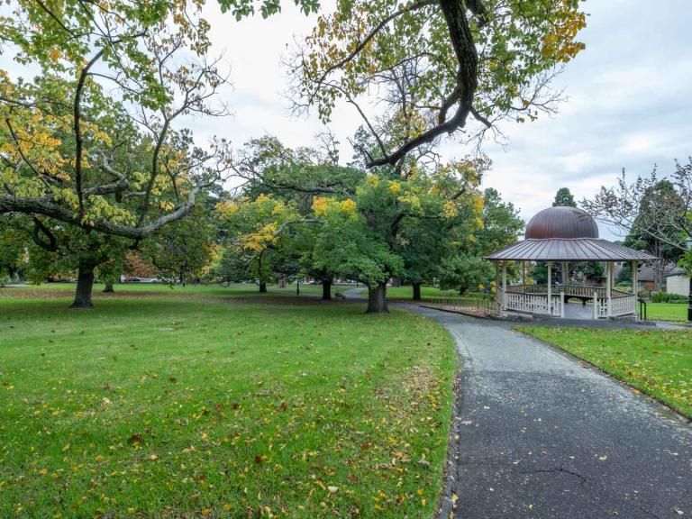 Large grass area with a rotunda and walking path to the right. There are scattered medium-sized trees and fallen leaves on the ground.