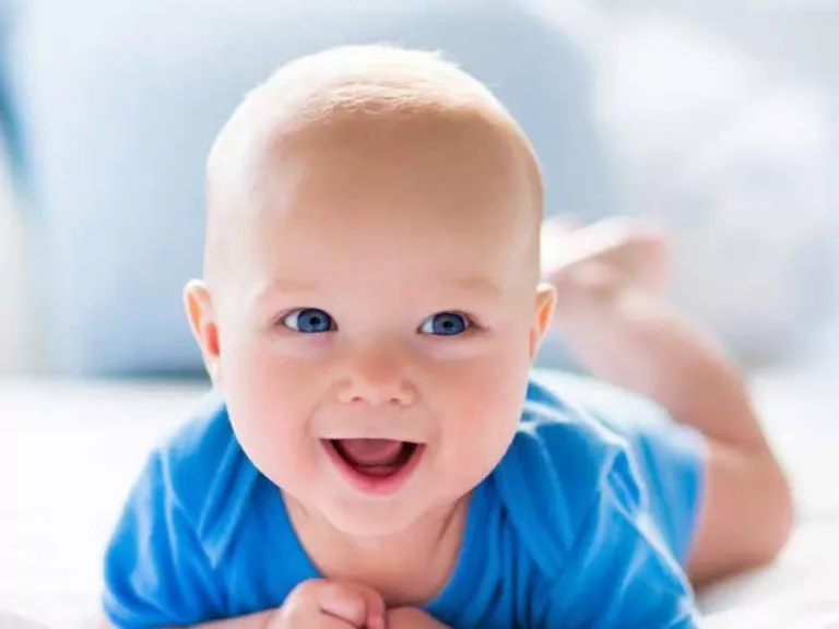 A baby laying on their stomach smiling and wearing a blue shirt