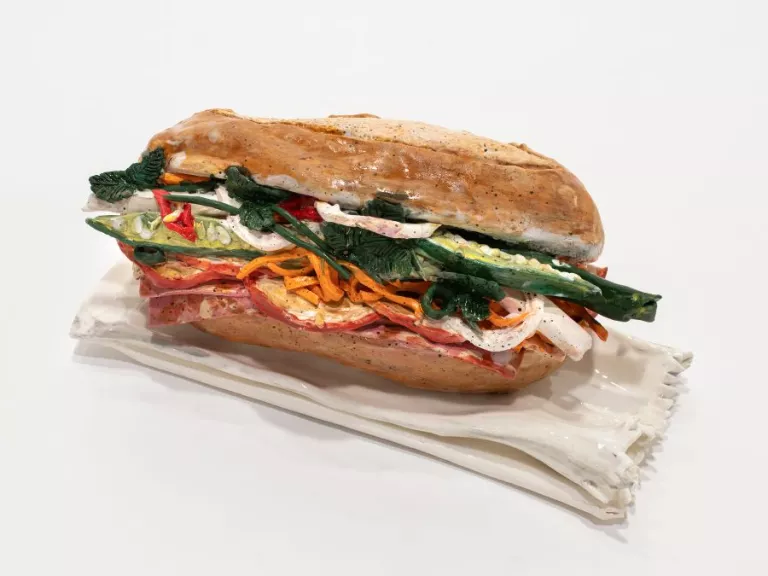 Image of ceramic Bánh mì sculpture on a white background. Some ingredients are identifiable as shredded carrot, chilli, coriander, salami, onion, and cucumber.  