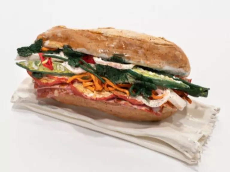 Image of ceramic Bánh mì sculpture on a white background. Some ingredients are identifiable as shredded carrot, chilli, coriander, salami, onion, and cucumber.  