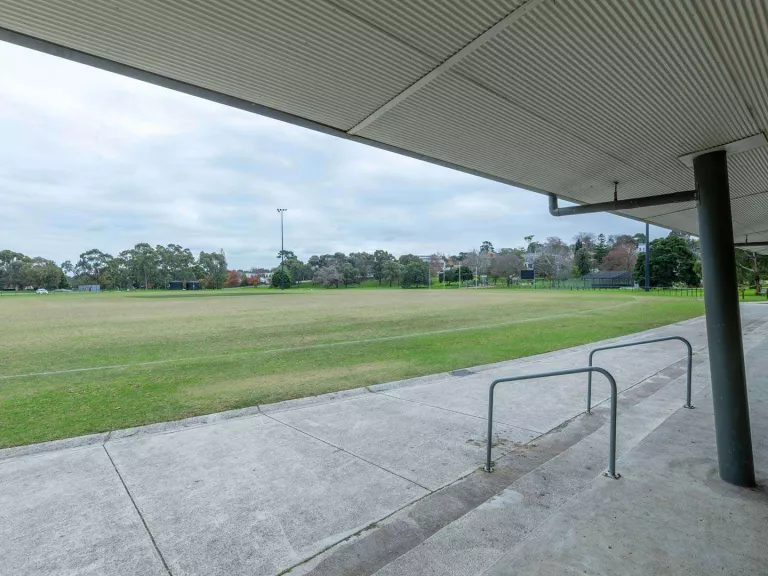 Looking out towards a green sports field from a sports grandstand . Trees and houses are visible in the background. 