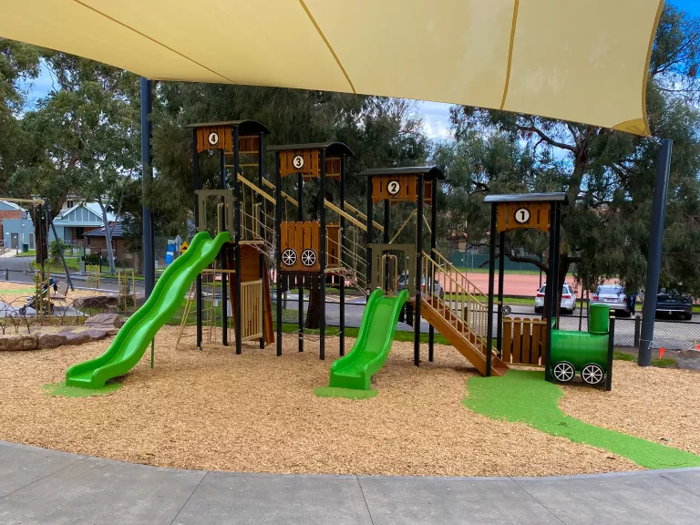 A children's playground has a timber climbing frame with green slides beneath a shade sail