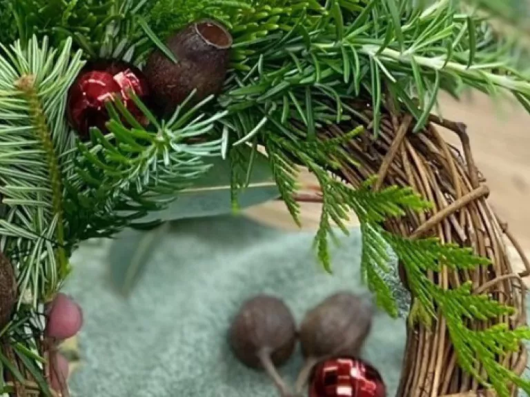 A hand is holding a wreath which is being made of green foliage and gumnuts 