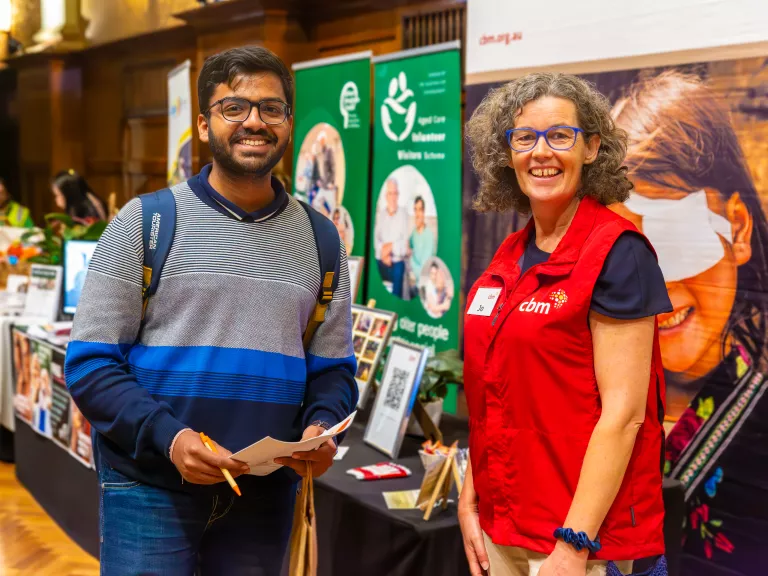 2 people standing at a volunteer expo booth. one is holding a pencil and paper.