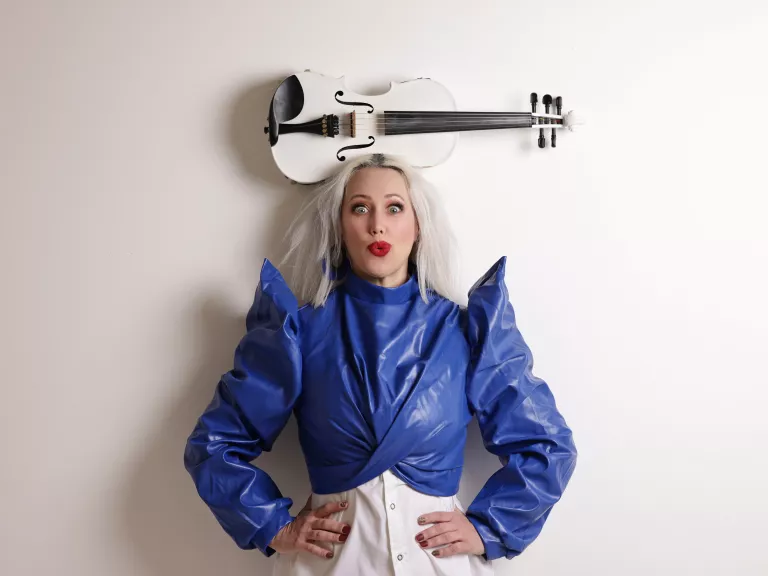 Woman with bleach blonde hair wearing a vibrant blue jacket with pointed shoulders balances a white violin on her head.