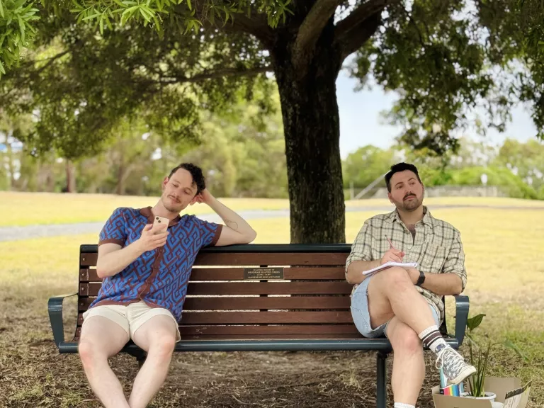 two people sitting on a park bench under a tree. One is looking at their phone while the other has a pen and paper