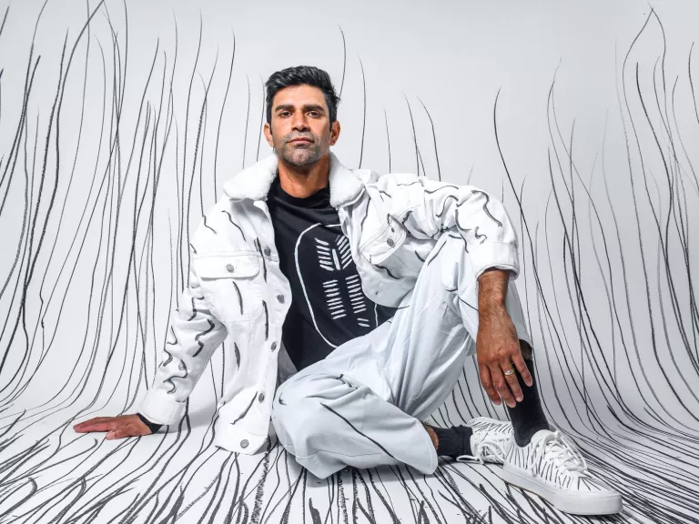 first nations man wearing white pants and jacket sits against a white backdrop with charcoal lines drawn from the floor up