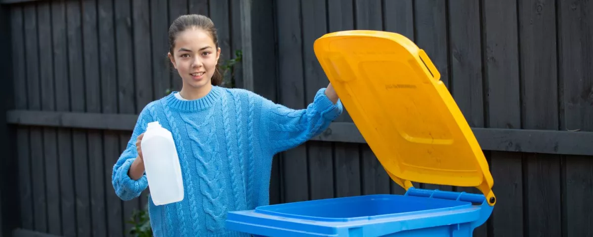 A young person standing next to an open recycling bin holding an empty milk jug