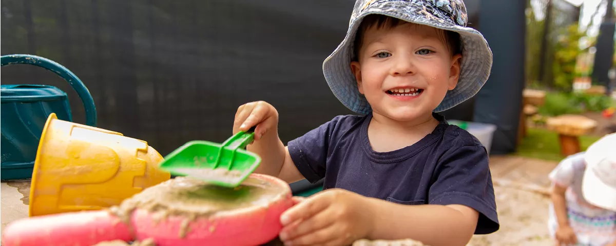 Young child in blue patterned hat plays with green spade in sandpit.