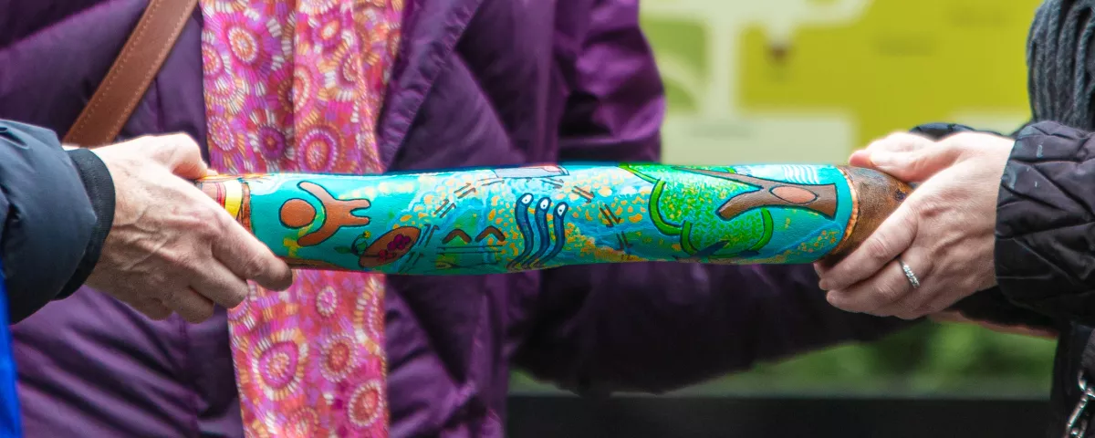 Painted aboriginal instrument being held at each end by a pair of hands