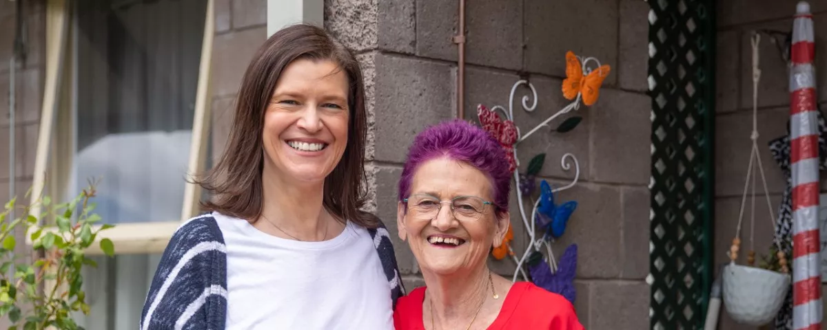 Two women side by side smiling at the camera and standing in front of a house