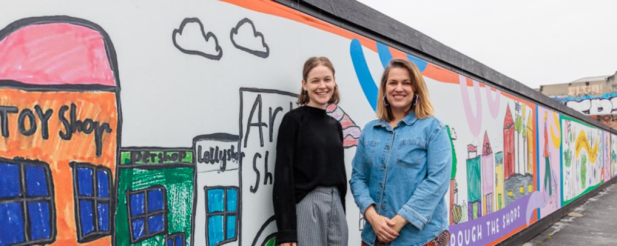 artists Angharad Neal-Williams and Nicole van Dijk stand in front of a section of the ‘Hart of Melbourne’ mural, which depicts shops and places in Hartwell drawn by school children