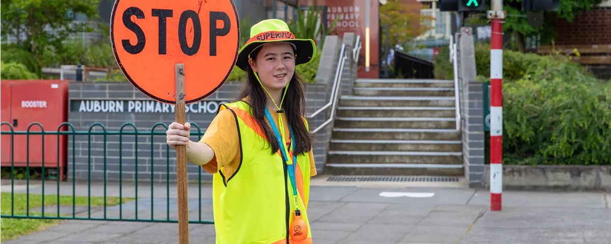 School crossing supervisor wearing hi-vis hat and vest holds stop sign in front of steps leading to Auburn Primary School.