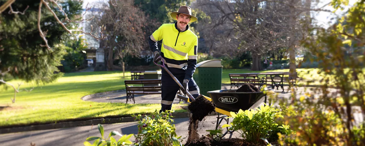 Gardener wearing hi-vis top and hat shovels compost from a wheelbarrow to a garden bed in a park.