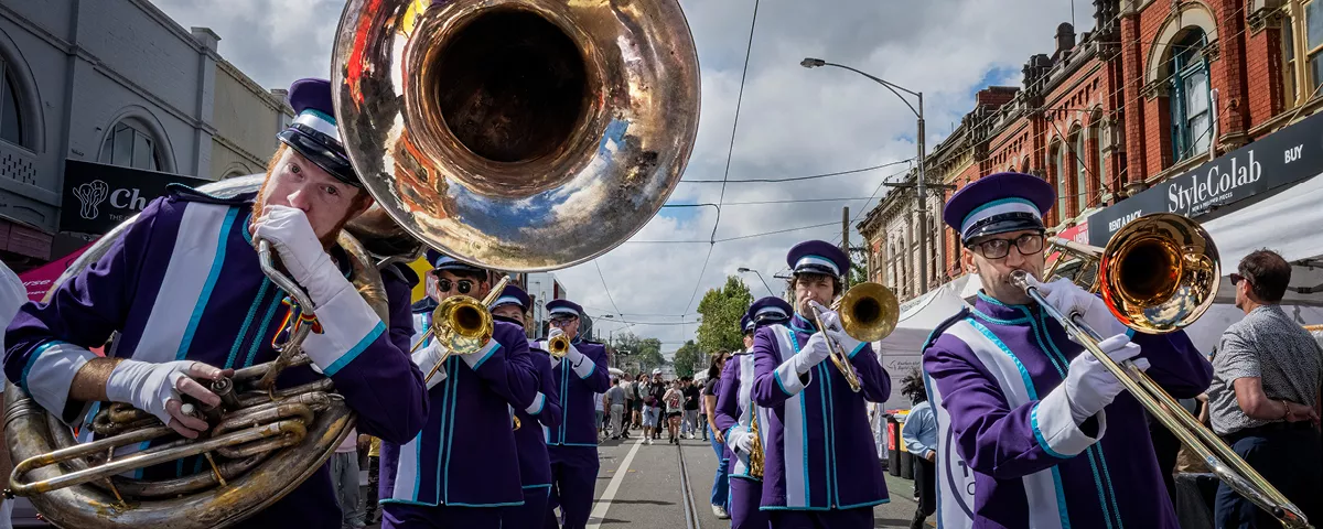 Members of brass band wearing purple and white uniforms walk along Glenferrie Road. 