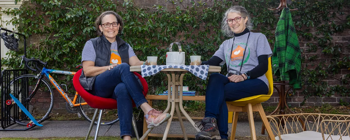 Two volunteers sit in garden beside table. In background are a bike, a coat rack, jacket and basket.