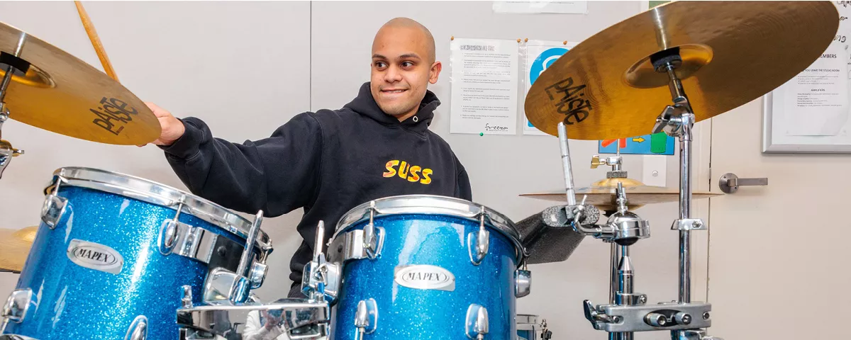 Smiling student wearing blue hooded jumper sits behind drum set playing blue drums and brass cymbals.