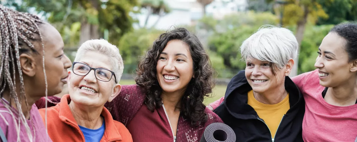 Five women are standing outside and smiling. The women are all wearing casual clothes and are standing with their arms around each other. They are all different ages and ethnicities.