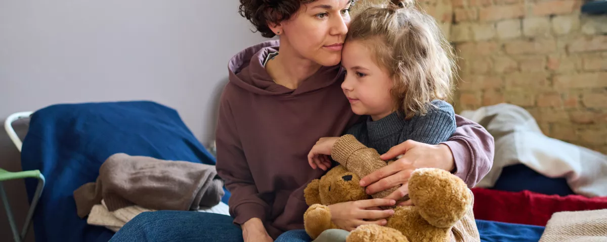 A woman sits on a bed, hugging a young girl who is holding a teddy bear. The woman’s hand rests on the girl's back in a comforting manner. There are blankets on the bed in the background.