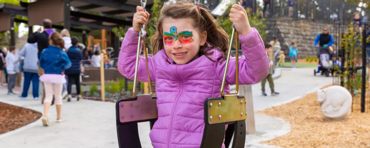 a child with colourful face paint is on the swings at a playground