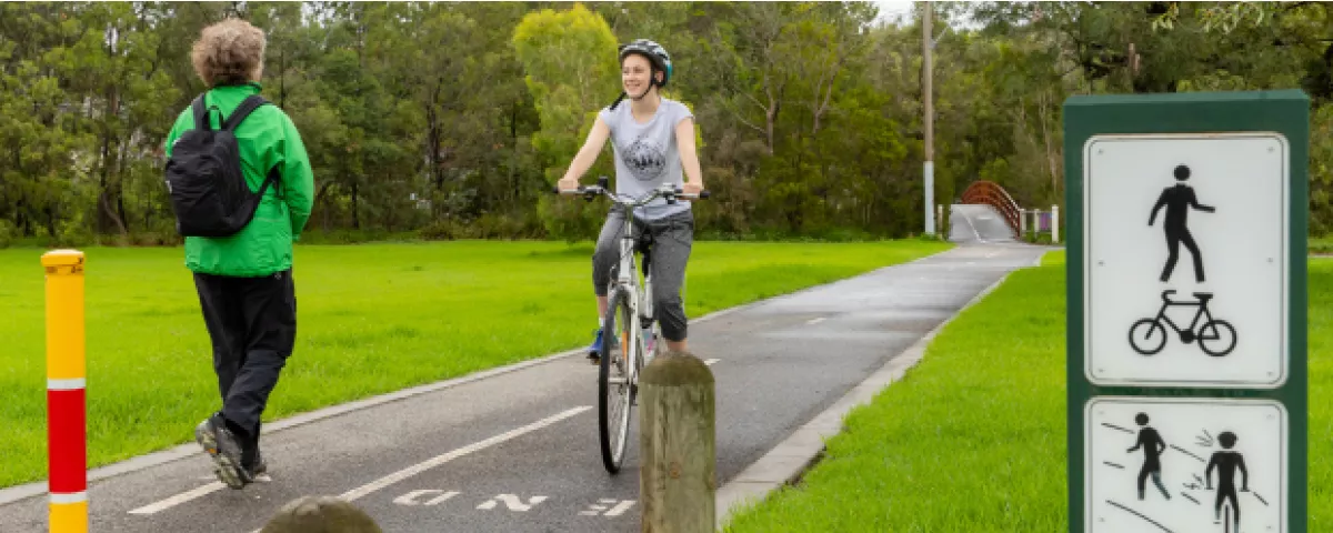 Cyclist on shared path approaches a walker moving the other way. Shared path sign in foreground shows figures cycling and walking.