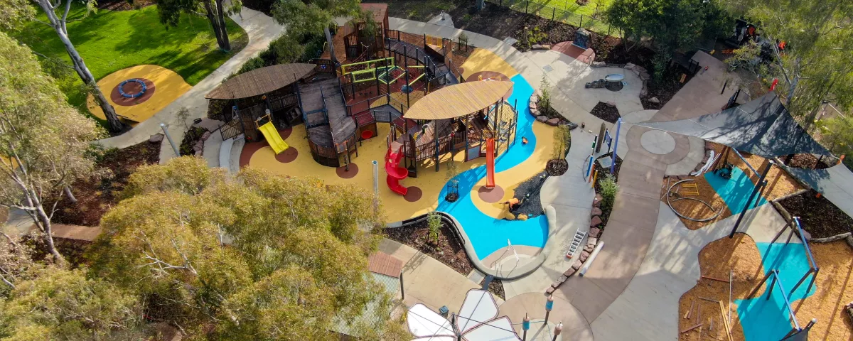 Drone image showing playground with timber play structures, play equipment and butterfly shade surrounded by parkland and trees