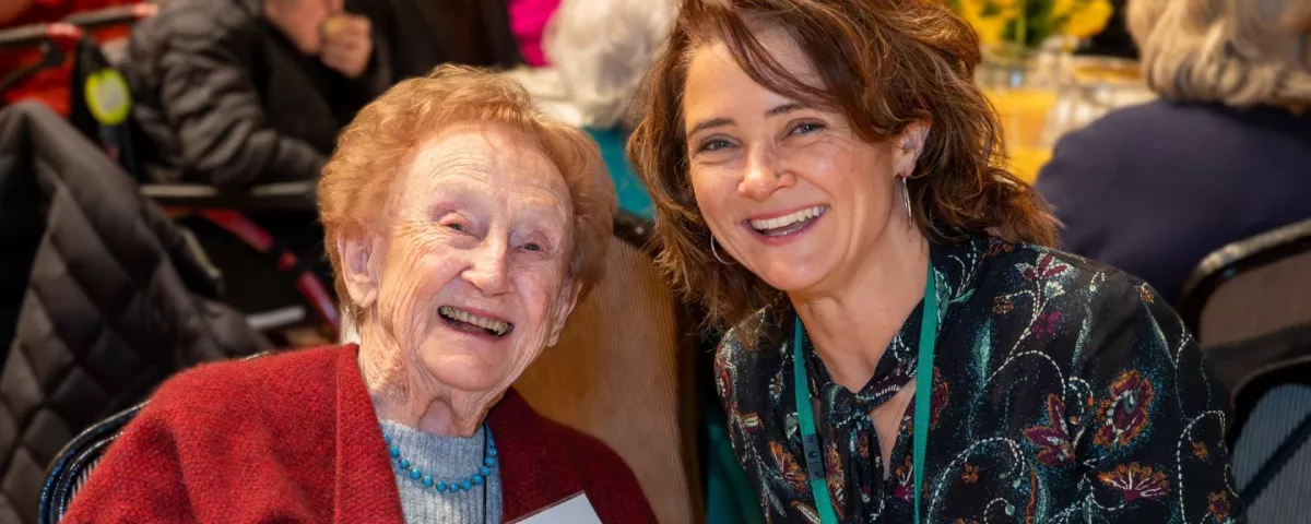 A senior citizen with a younger person seated in a room with banquet tables