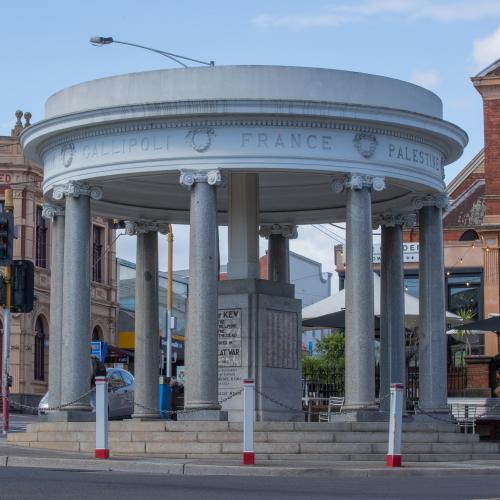 Street view of a cenotaph building on the edge of a roundabout with a historic building in background