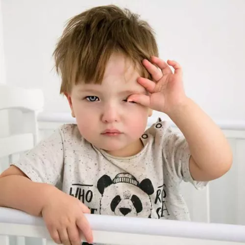 A toddler rubbing their eyes looking tired while standing in a cot