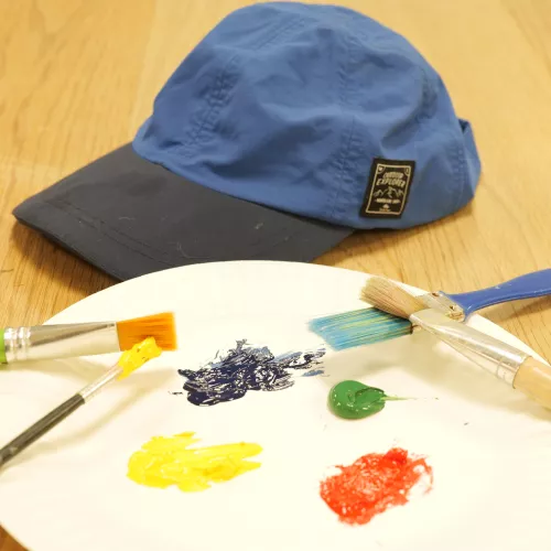 A paint palette with paints and paint brushes next to a black and blue baseball cap on a wooden table. 