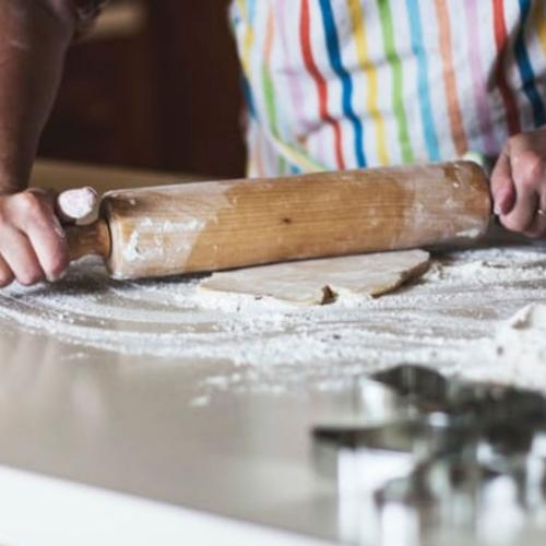 photo of a person rolling dough with a rolling pin. They are wearing a striped apron and there is lots of flour on the bench.