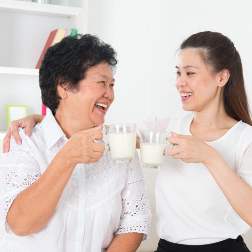 2 Chinese women wearing white shirts are holding glass mugs of milk and smiling at each other