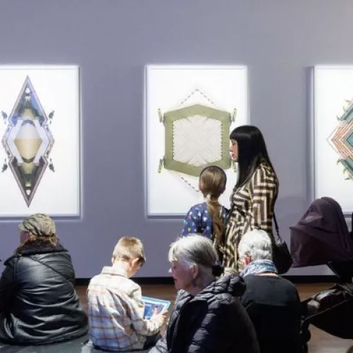 Image of man in gallery speaking to a crowd of people. Artwork is hung on wall in background. 