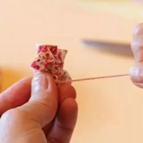 Photo of hands tying a pink piece of yarn around a small bunched up piece of pink patterned fabric. A blurry fabric flower is positioned in the background behind the hands.