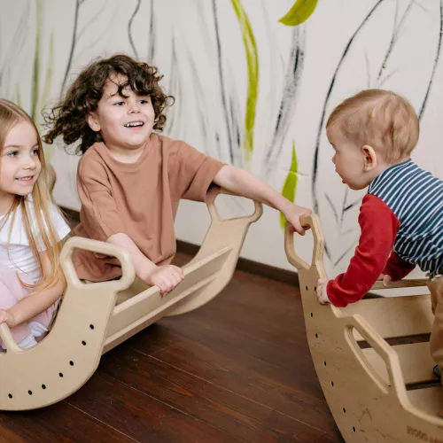 Three young children are sitting in toy rockers smiling. Two children are in one rocker looking at and smiling at the other child in the other rocker.