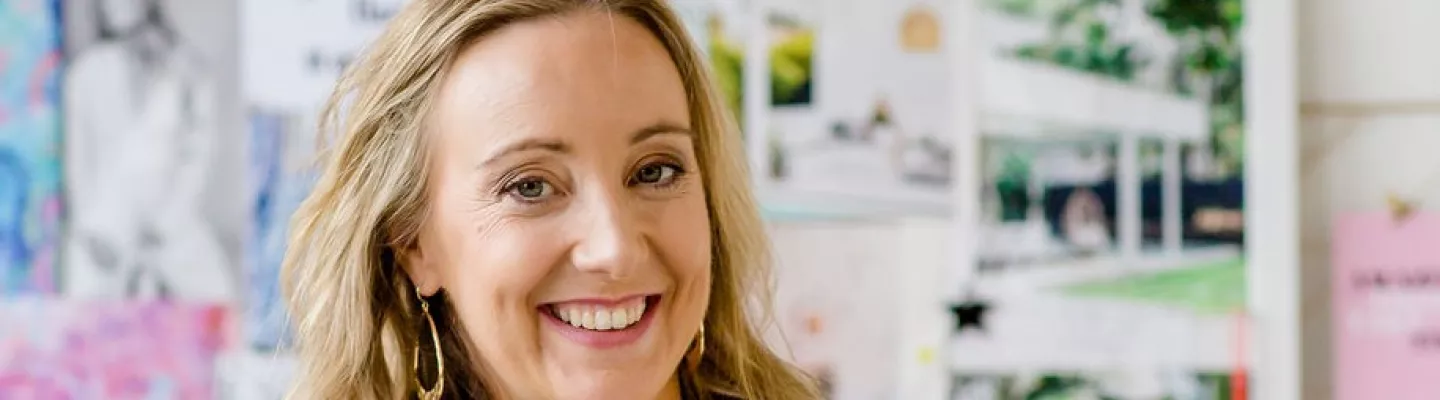 Smiling woman in a navy blazer with a colourful pinboard behind her