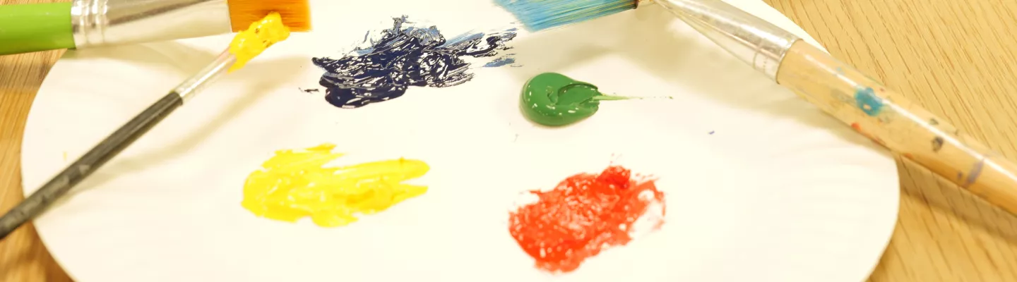 A paint palette with paints and paint brushes next to a black and blue baseball cap on a wooden table. 