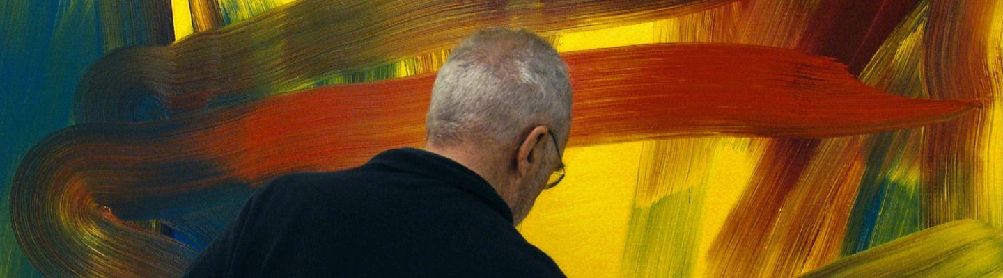 Photo of man with grey hair and black jacket standing with his back turned in front of an abstract blue, yellow and red painting. 