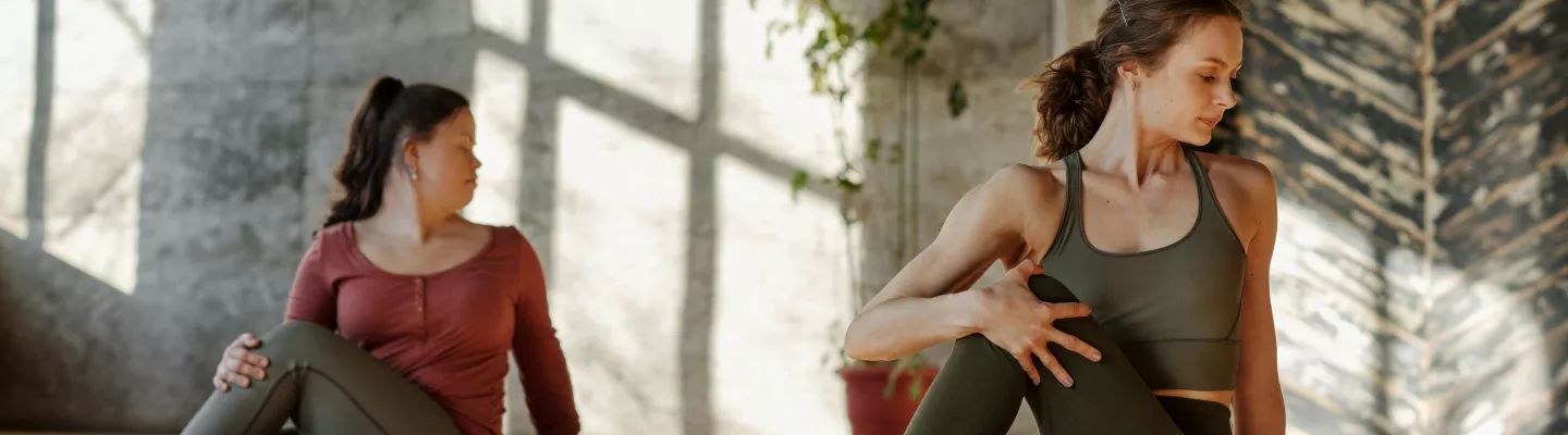 Two females in exercise outfits are sitting on yoga mats doing the same stretching exercise. In the background there are sun shadows on the wall.