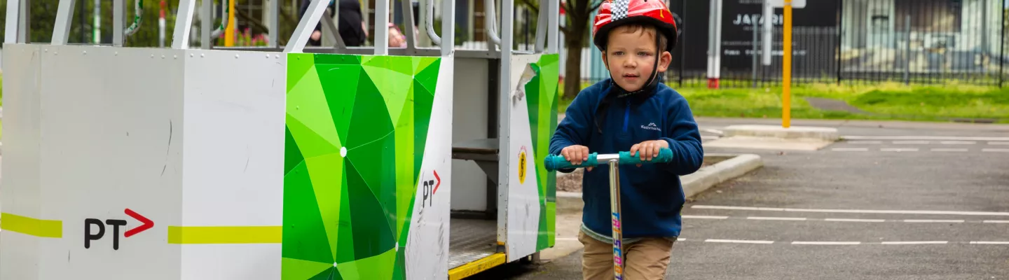 Child wearing a red helmet on a scooter rides past a small scale tram