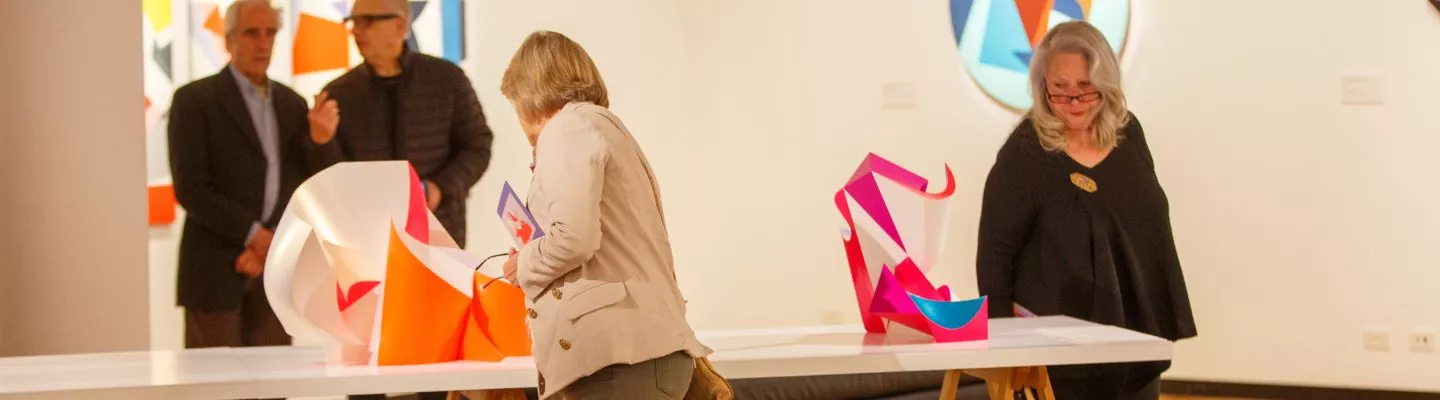 Visitors in an art gallery viewing colourful abstract sculptures sitting on tables, with geometric artworks on the walls behind them.