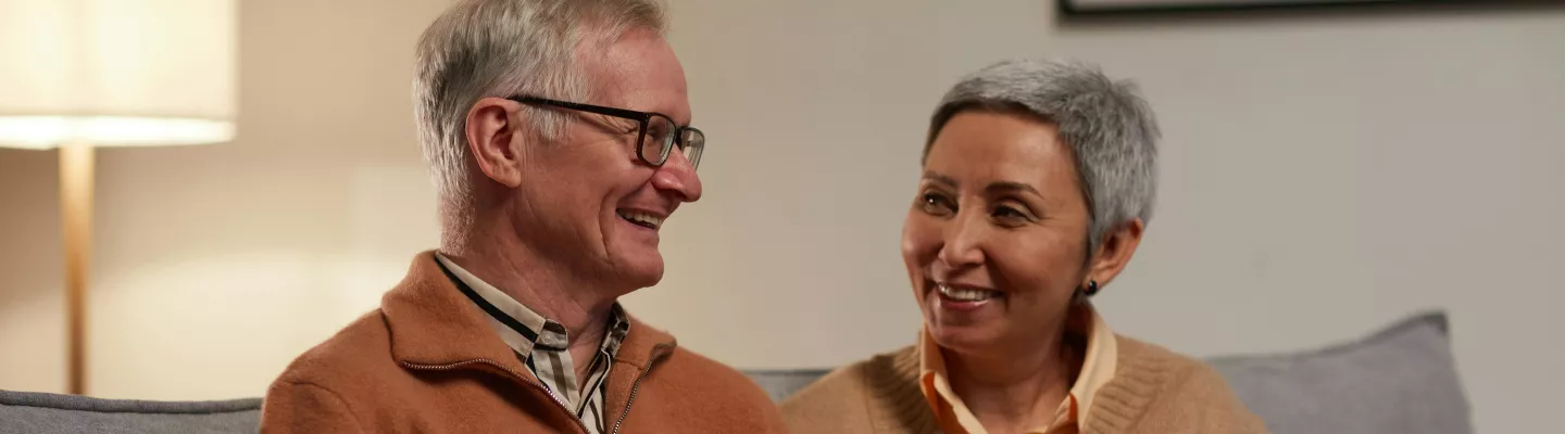 A man and woman sitting on a couch looking at each other smiling. The man has a laptop on his lap.