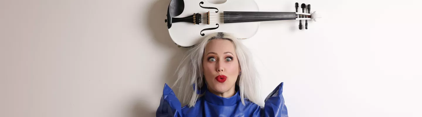 Woman with bleach blonde hair wearing a vibrant blue jacket with pointed shoulders balances a white violin on her head.