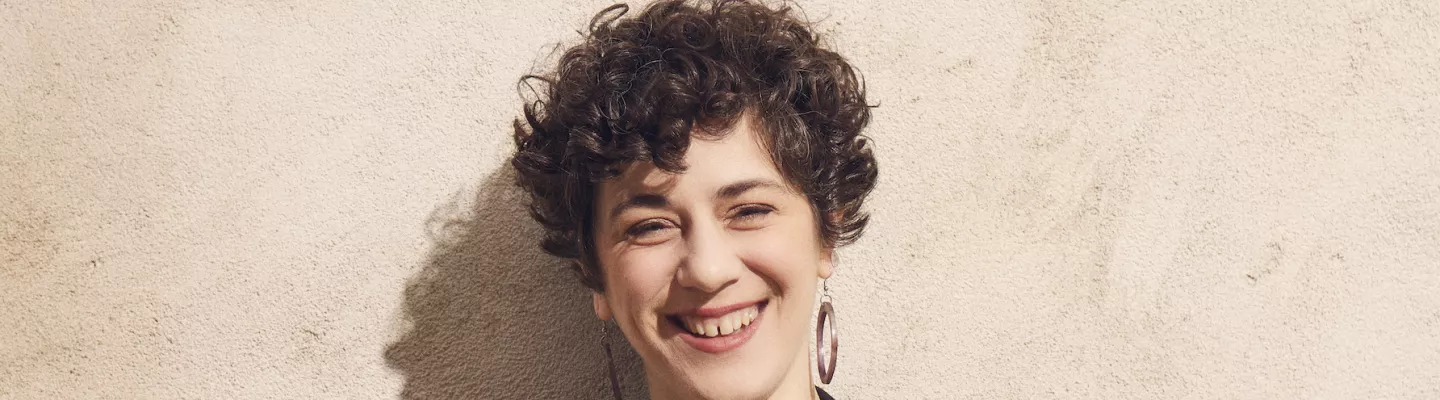 person with short curly hair wearing black standing against a brick wall