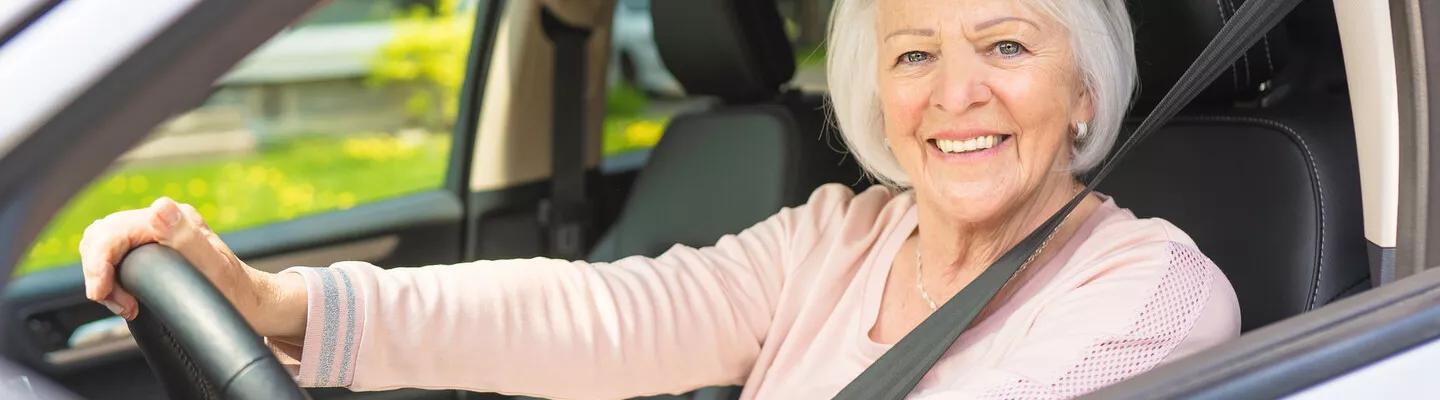 A woman is sitting in a car with her hands on the wheel smiling at the camera through the drivers side window. 