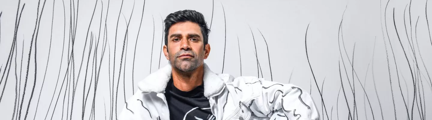 first nations man wearing white pants and jacket sits against a white backdrop with charcoal lines drawn from the floor up