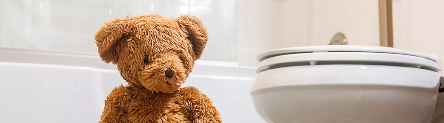 A toy teddy bear sits on a green potty, in a bathroom.