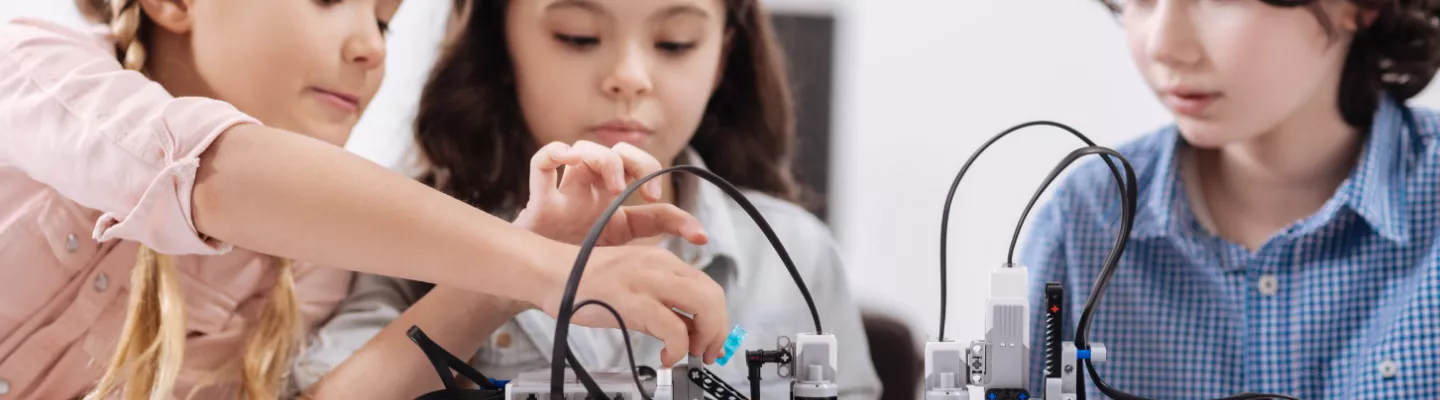 Three children are sitting and looking at a robotic lego construction. Two of the children are touching the construction.