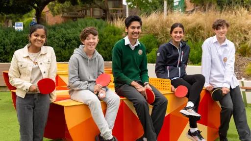 A group of 5 young people sitting on an outdoor table tennis table holding paddles.