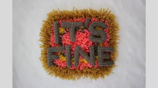 A piece of carpet with brown, pink, yellow, and grey wool in concentric cicles, overlaid with crocheted letters spelling out 'It's fine'